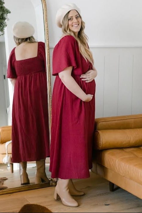 woman wearing a burgundy maternity dress with flutter sleeves, holding her baby bump while standing next to a mirror