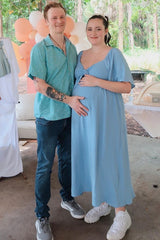 woman wearing a light blue maternity dress and cradling her baby bump, standing next to a man in a patterned shirt