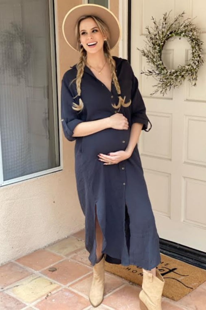 Smiling pregnant woman in a navy blue button-down midi dress and tan hat holding her baby bump on a porch