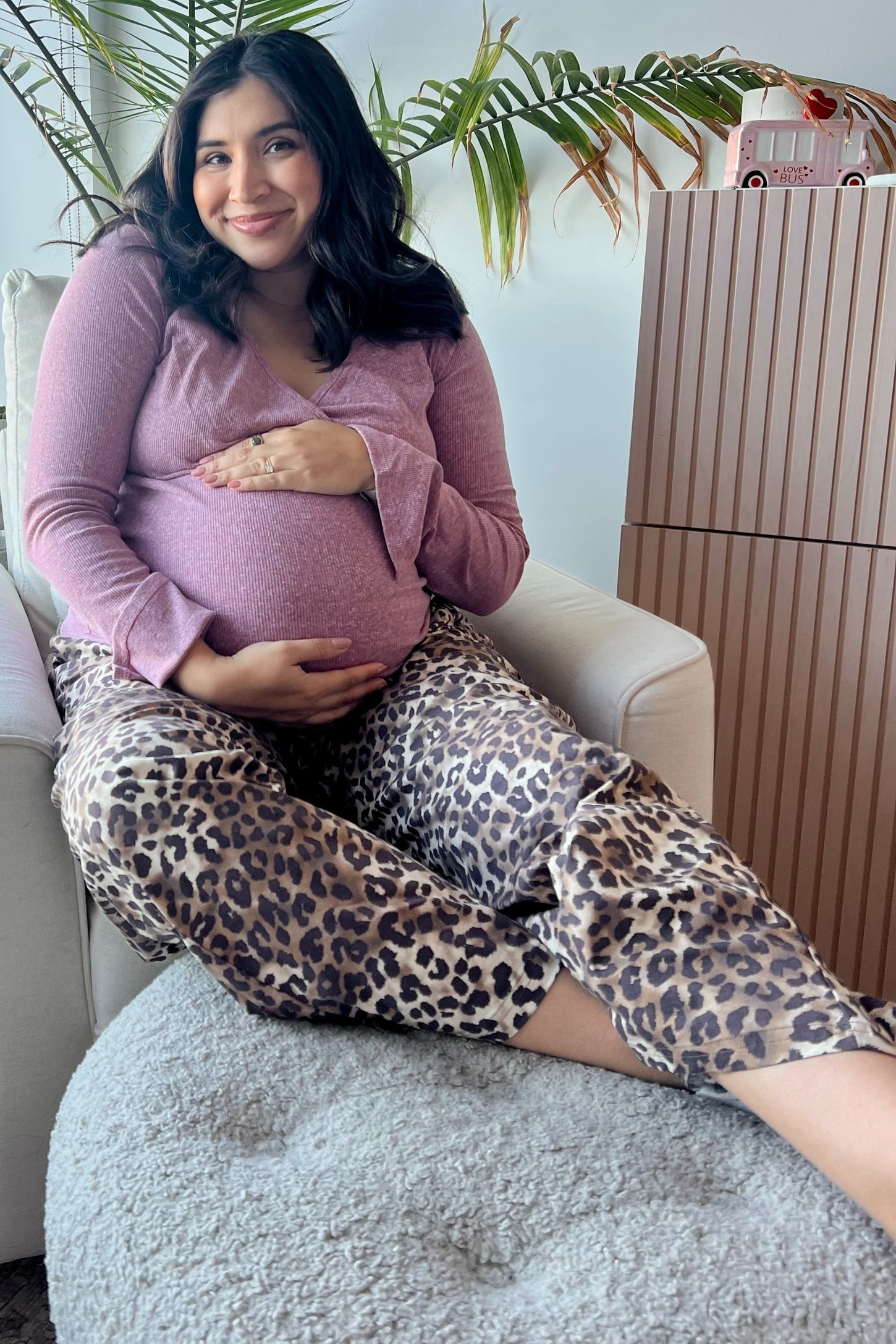 woman wearing a light purple long sleeve maternity top and leopard print maternity pants, sitting and holding her baby bump