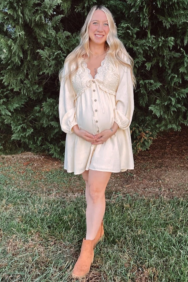 woman wearing a cream colored maternity dress with lace details, smiling and holding her baby bump outdoors