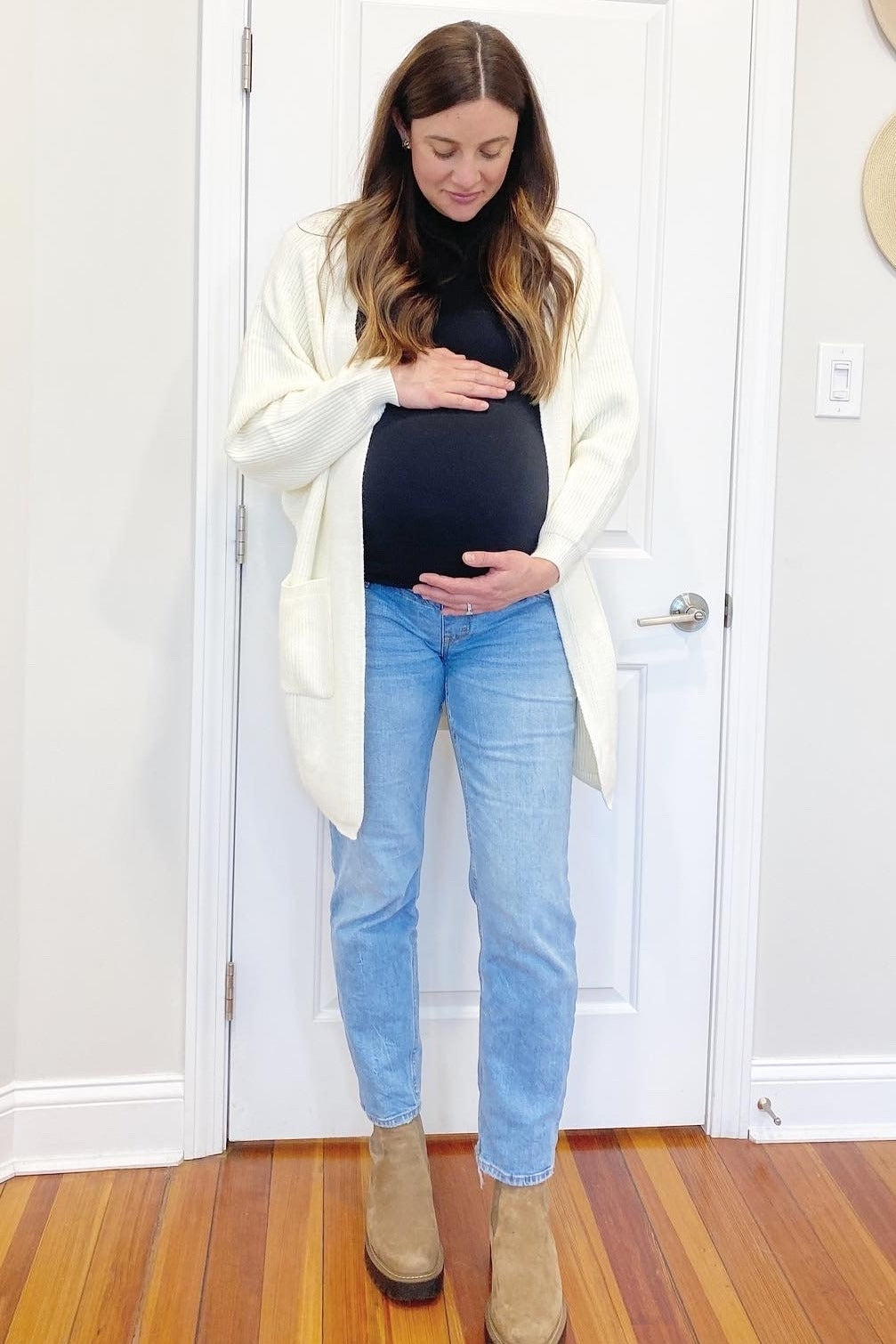 woman wearing a black turtleneck and cream cardigan, showing her baby bump in light blue jeans and tan boots
