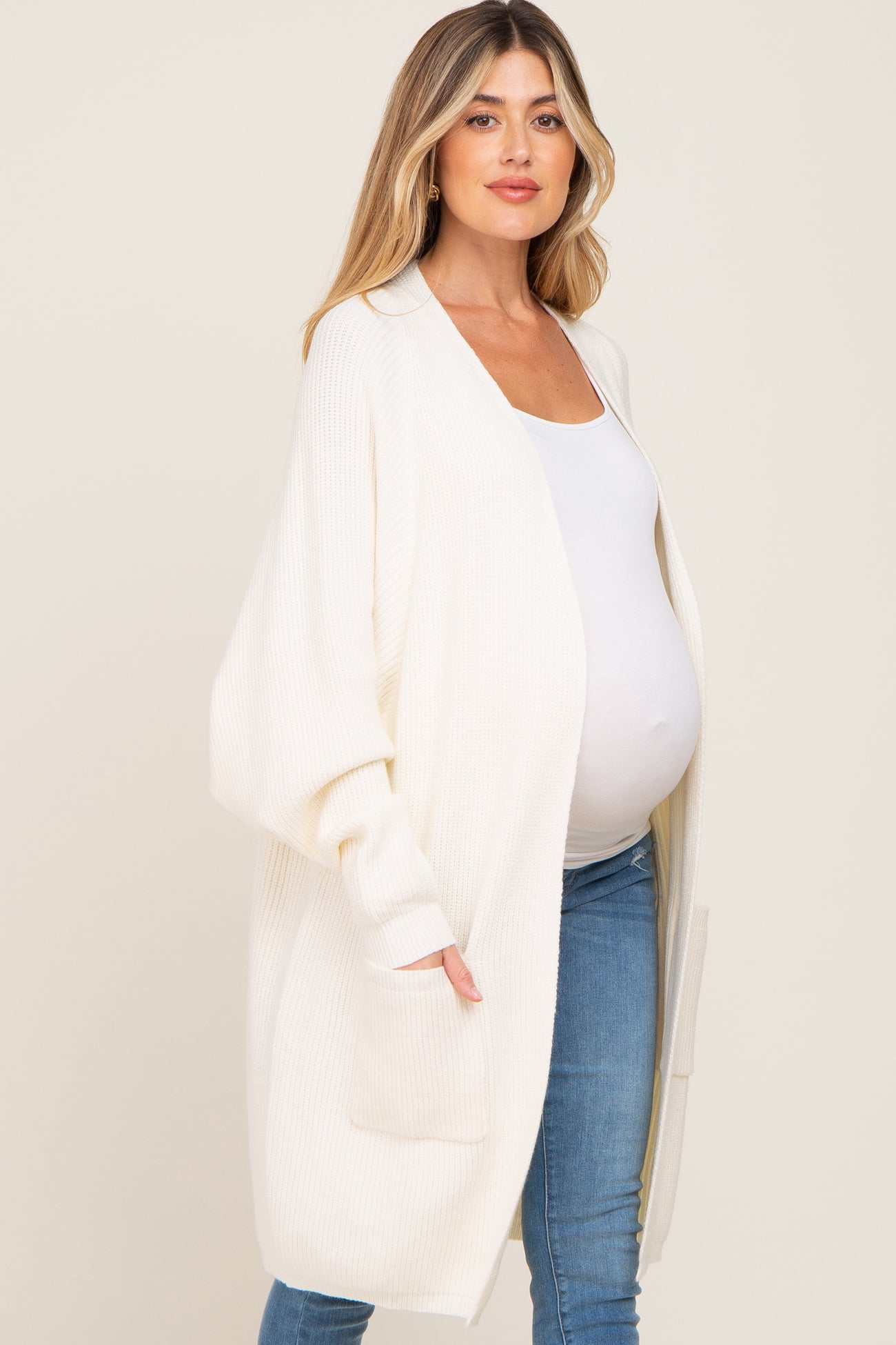 pregnant woman wearing a long white knit maternity cardigan over a white top and blue jeans