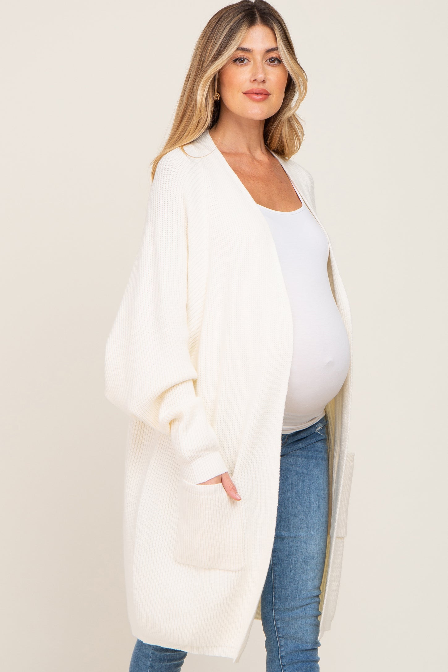pregnant woman wearing a long white knit maternity cardigan over a white top and blue jeans