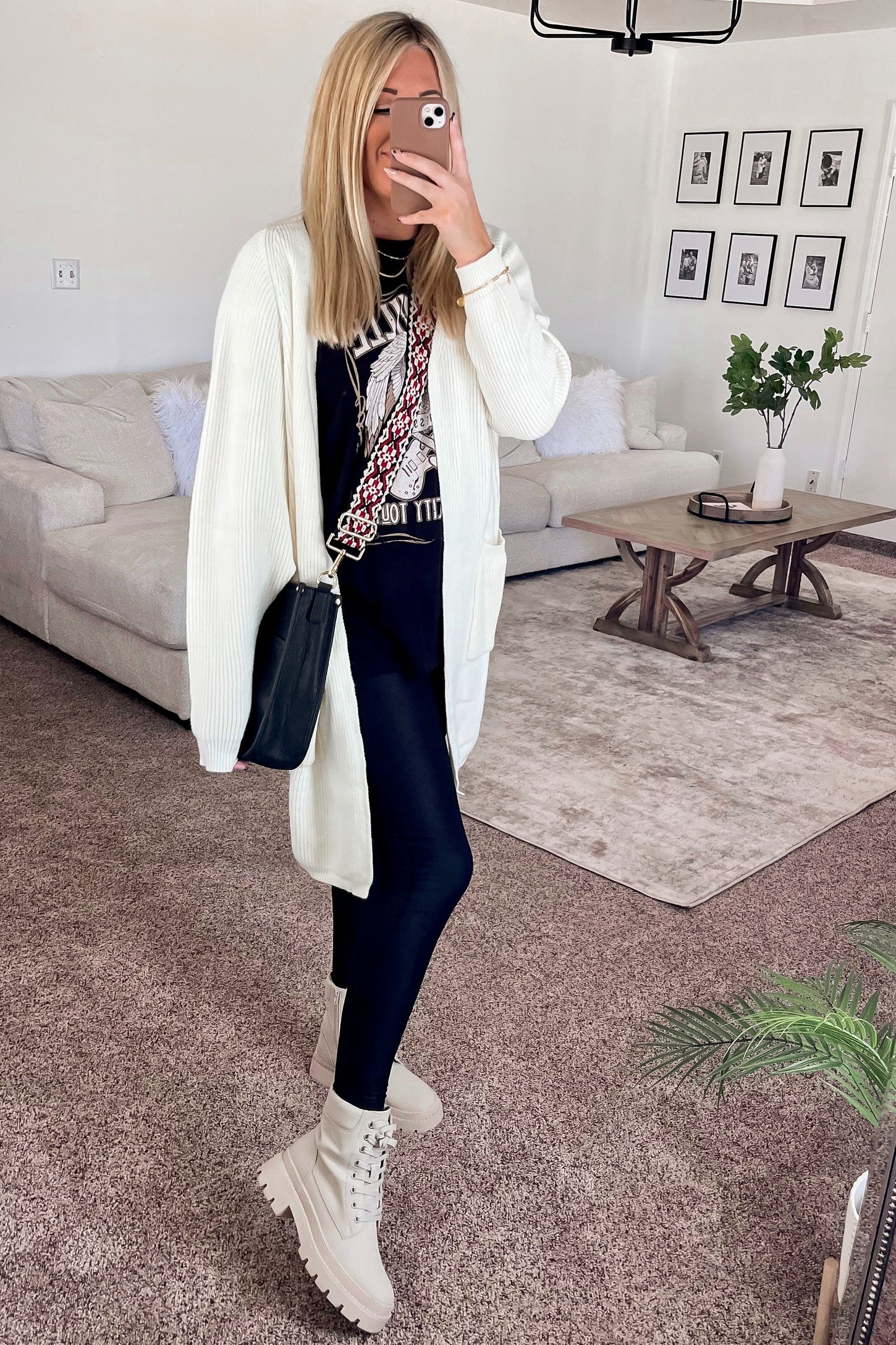 woman wearing a white long cardigan, black graphic t-shirt, black leggings, and beige combat boots, posing in a living room