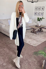 woman wearing a white long cardigan, black graphic t-shirt, black leggings, and beige combat boots, posing in a living room