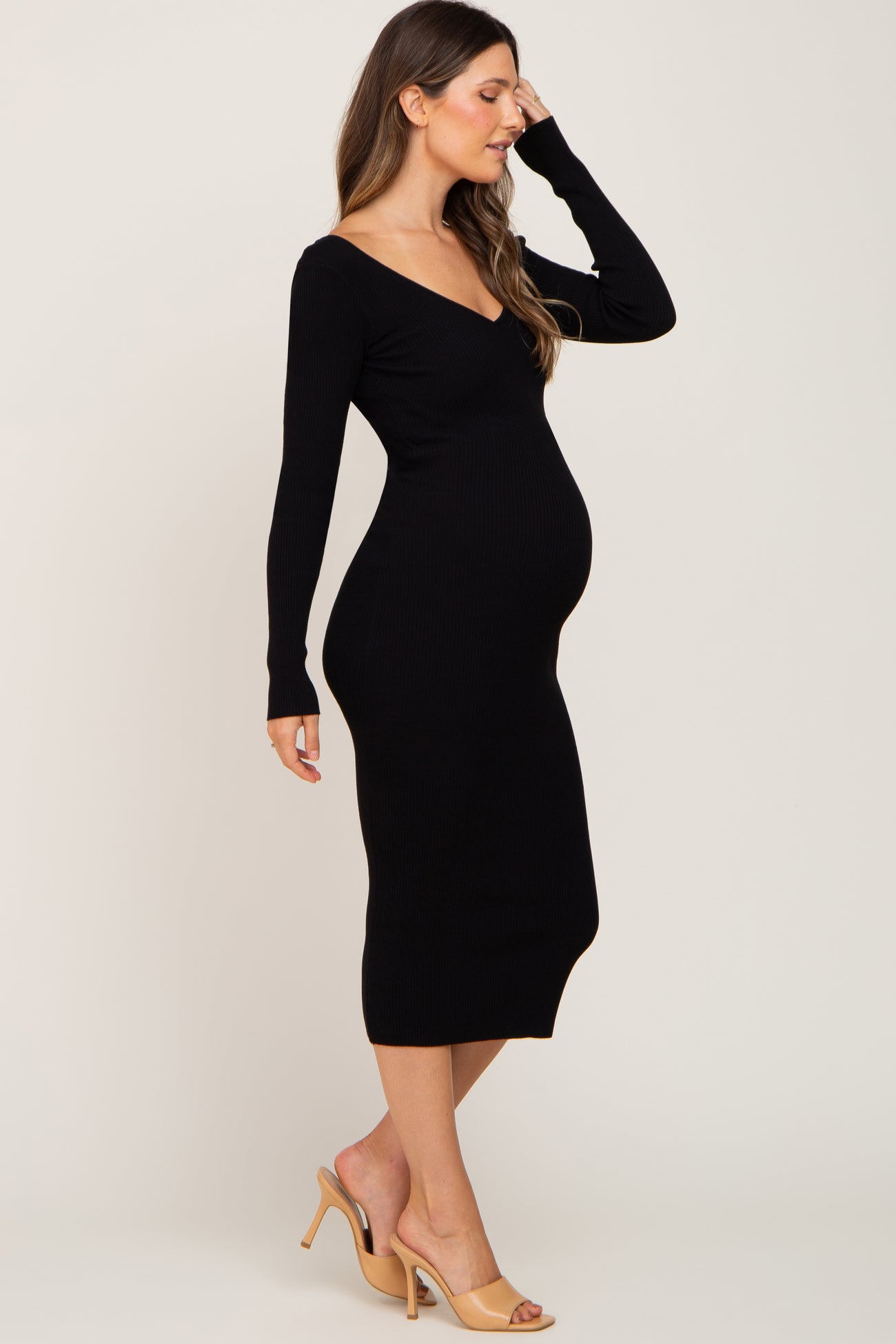 Pregnant woman wearing a black long-sleeve ribbed maternity midi dress and tan heels against a neutral background.