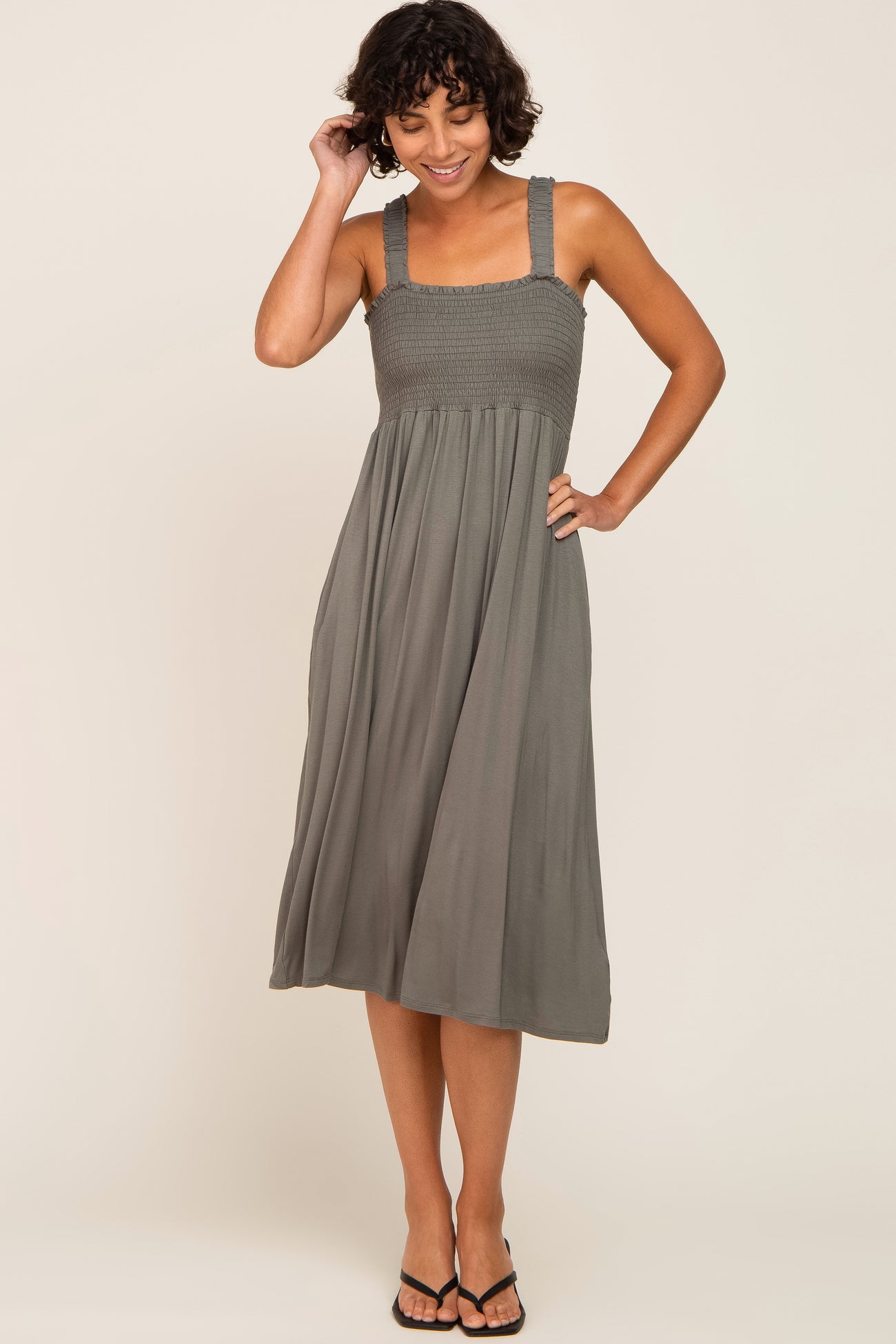 woman wearing a sage green midi dress with smocked bodice and wide adjustable straps, standing and smiling