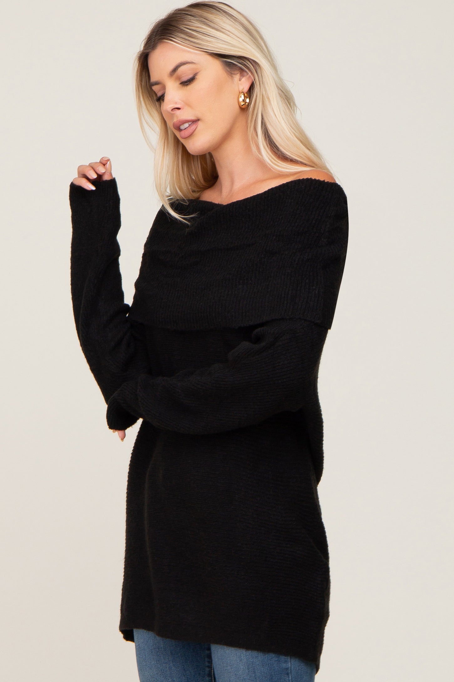 woman wearing a black off-the-shoulder knit sweater and blue jeans against a white background