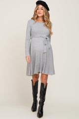 woman wearing a gray long sleeve maternity dress with a tie waist and black knee-high boots, showing her baby bump