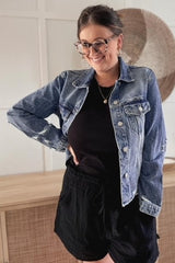woman wearing a distressed blue denim jacket over a black shirt and shorts with tortoise shell glasses