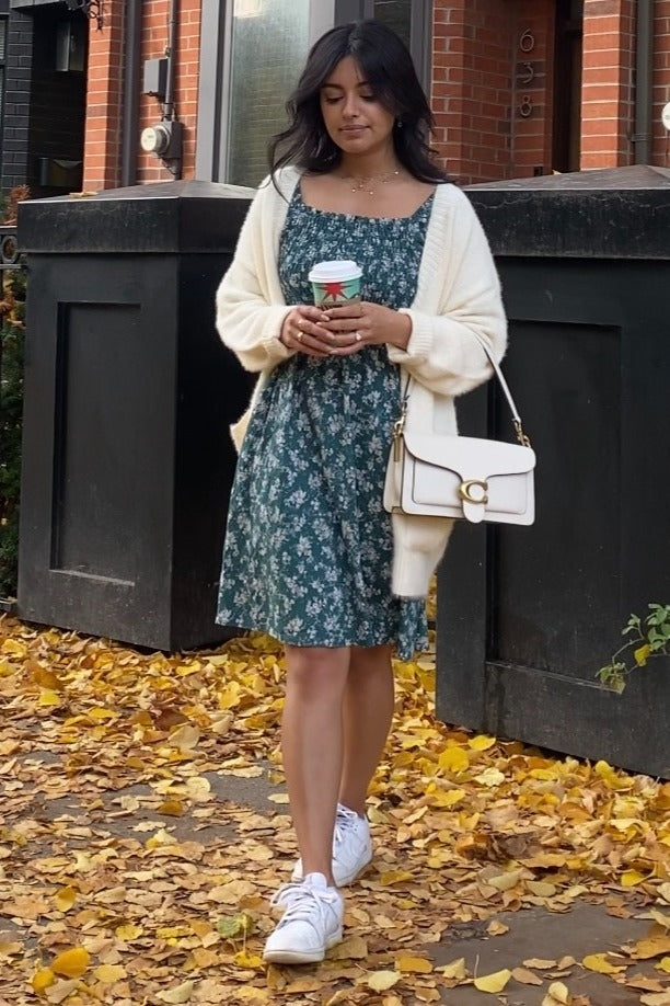 Woman in green floral dress and white cardigan holding coffee on autumn sidewalk with yellow leaves
