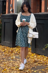 Woman in green floral dress and white cardigan holding coffee on autumn sidewalk with yellow leaves