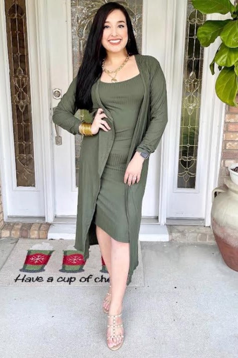 woman wearing an olive green fitted dress with a matching open cardigan and strappy heels standing by a door