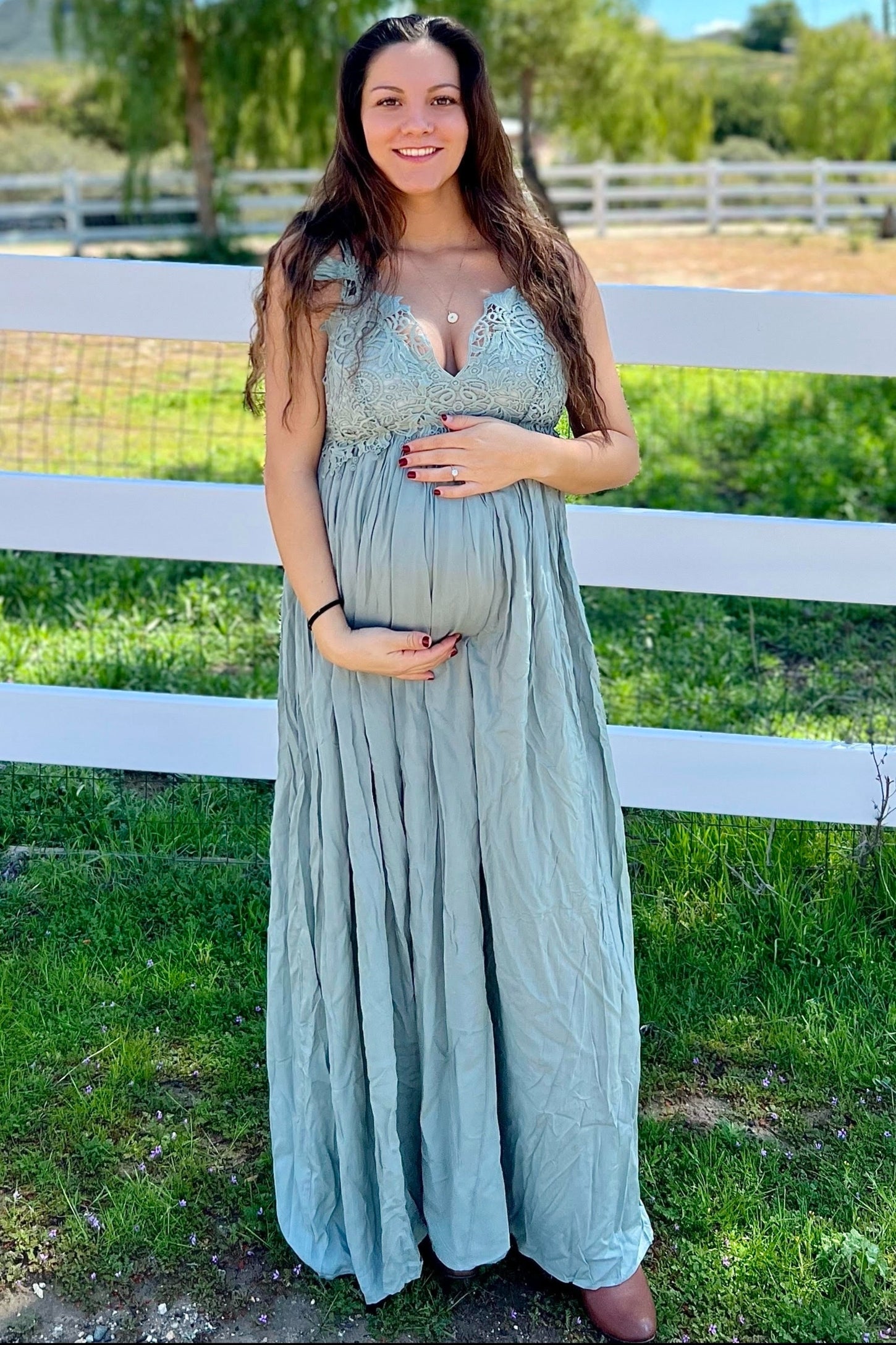 woman wearing a light green maternity dress with lace bodice and a visible baby bump, standing outdoors by a white fence