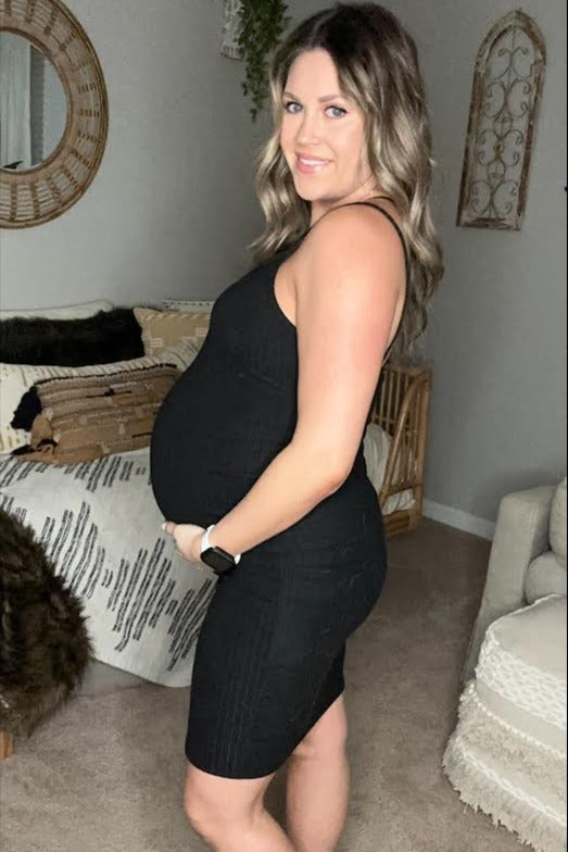 woman wearing a black ribbed maternity dress, showcasing her baby bump while posing in a cozy living room