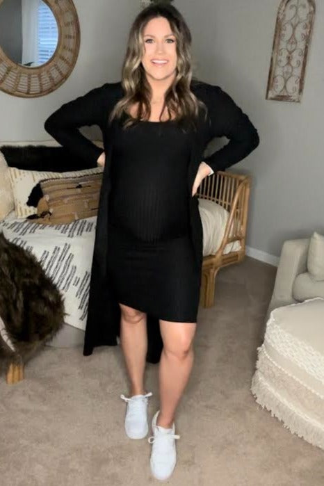 woman wearing a black maternity dress and long cardigan, showing her baby bump, standing in a cozy living room