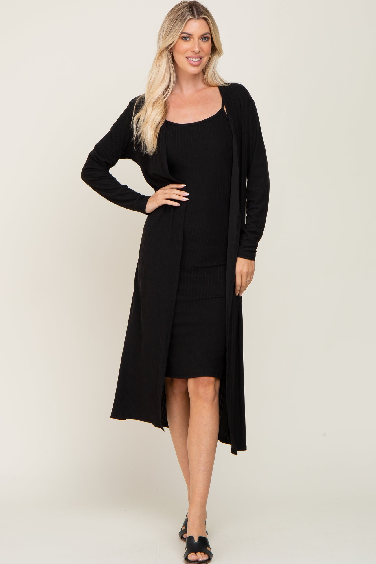 woman wearing a black long sleeve cardigan over a black fitted dress, standing with one hand on her hip