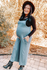 woman wearing a teal maternity jumpsuit with pockets and a black long sleeve top, displaying her baby bump