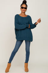woman wearing a teal oversized sweater, blue skinny jeans, and tan ankle boots, accessorized with sunglasses and earrings