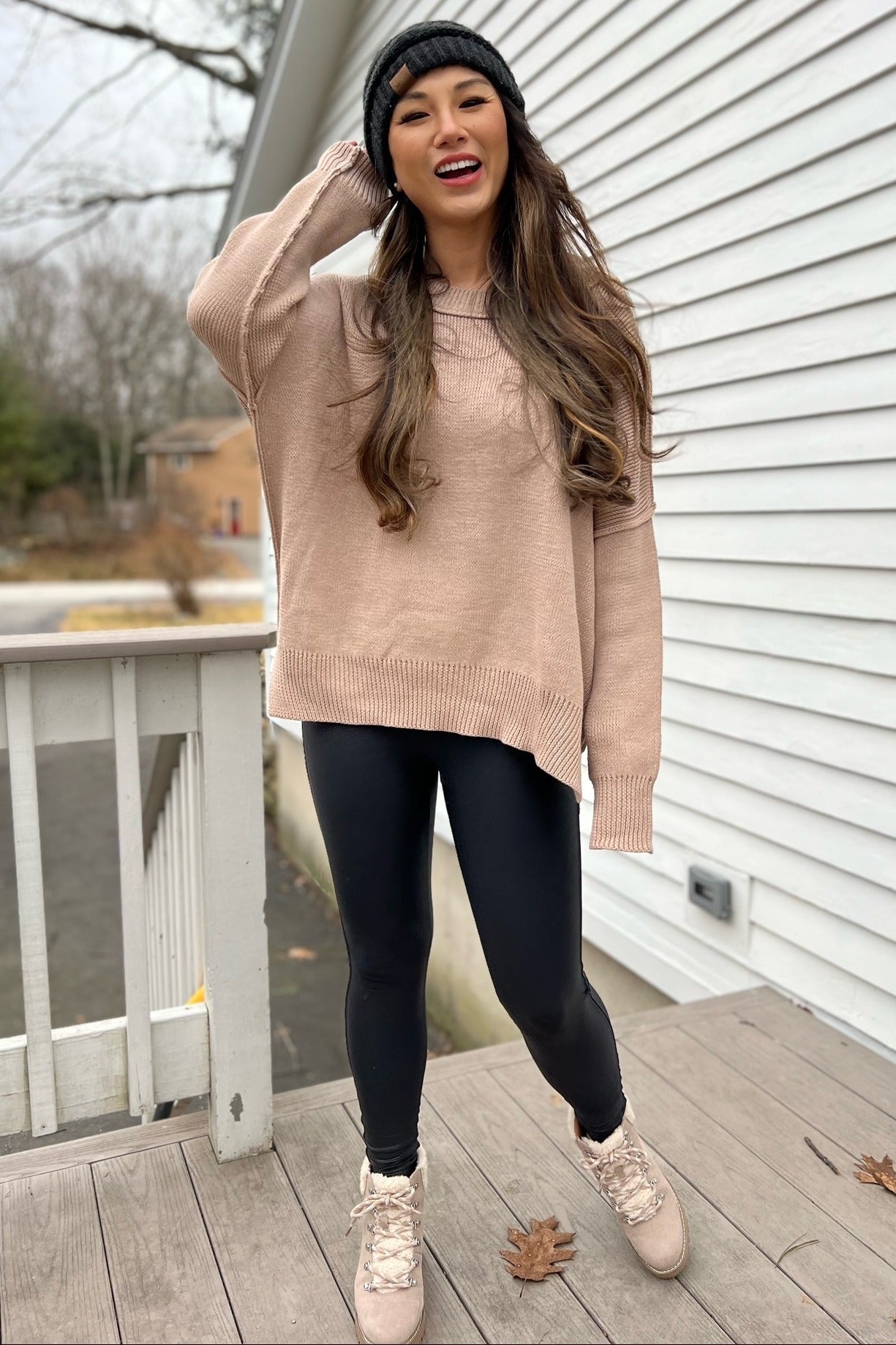 woman wearing a light pink oversized sweater, black leggings, and pink boots, smiling outdoors with a beanie on her head