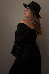 woman wearing a black off-the-shoulder maternity dress with flowing sleeves and a black hat, showcasing her baby bump