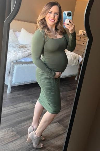 pregnant woman in olive green long sleeve maternity dress taking mirror selfie in bedroom