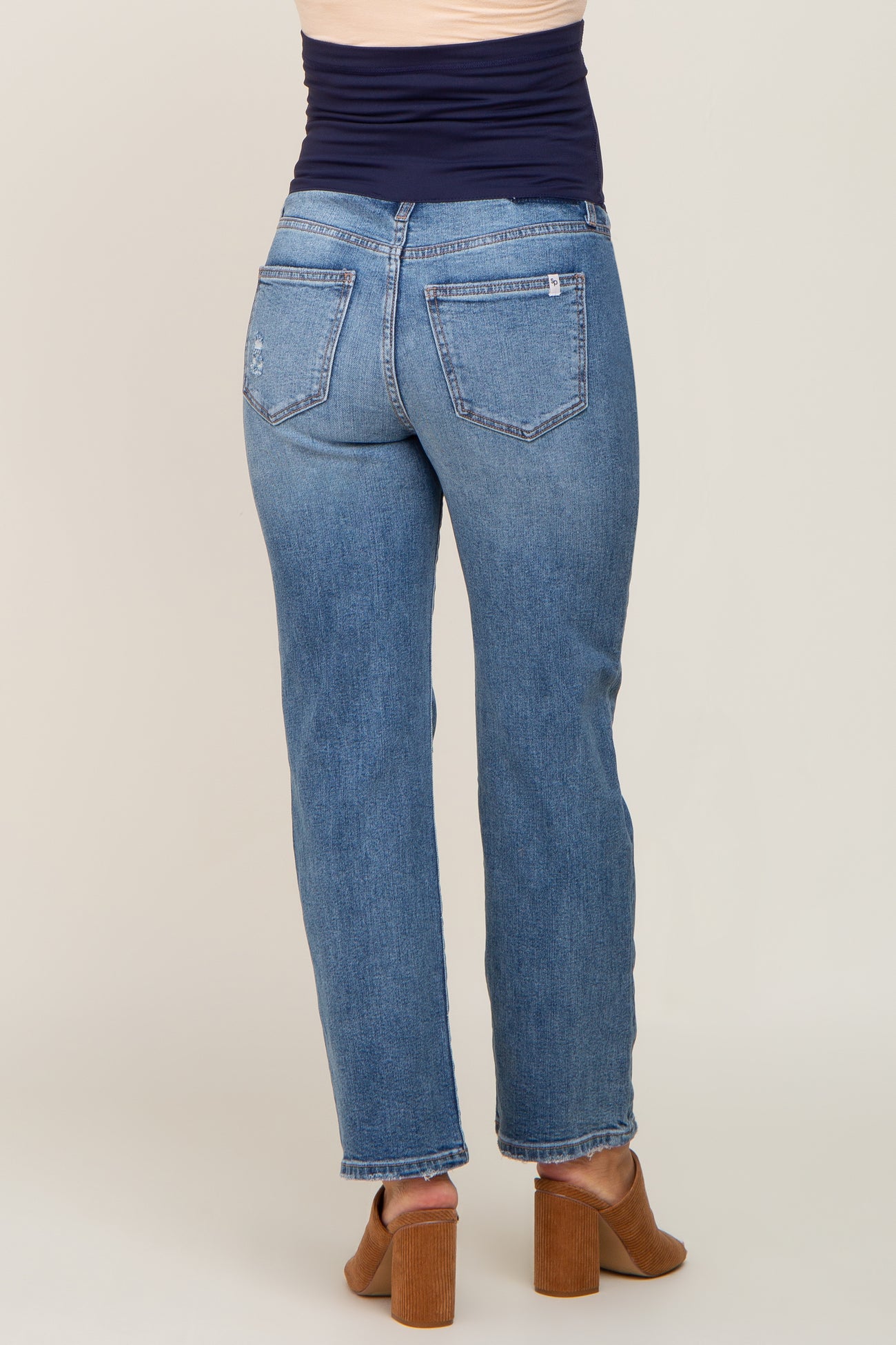 Back view of woman wearing blue straight leg maternity jeans with a navy belly band and brown heeled mules