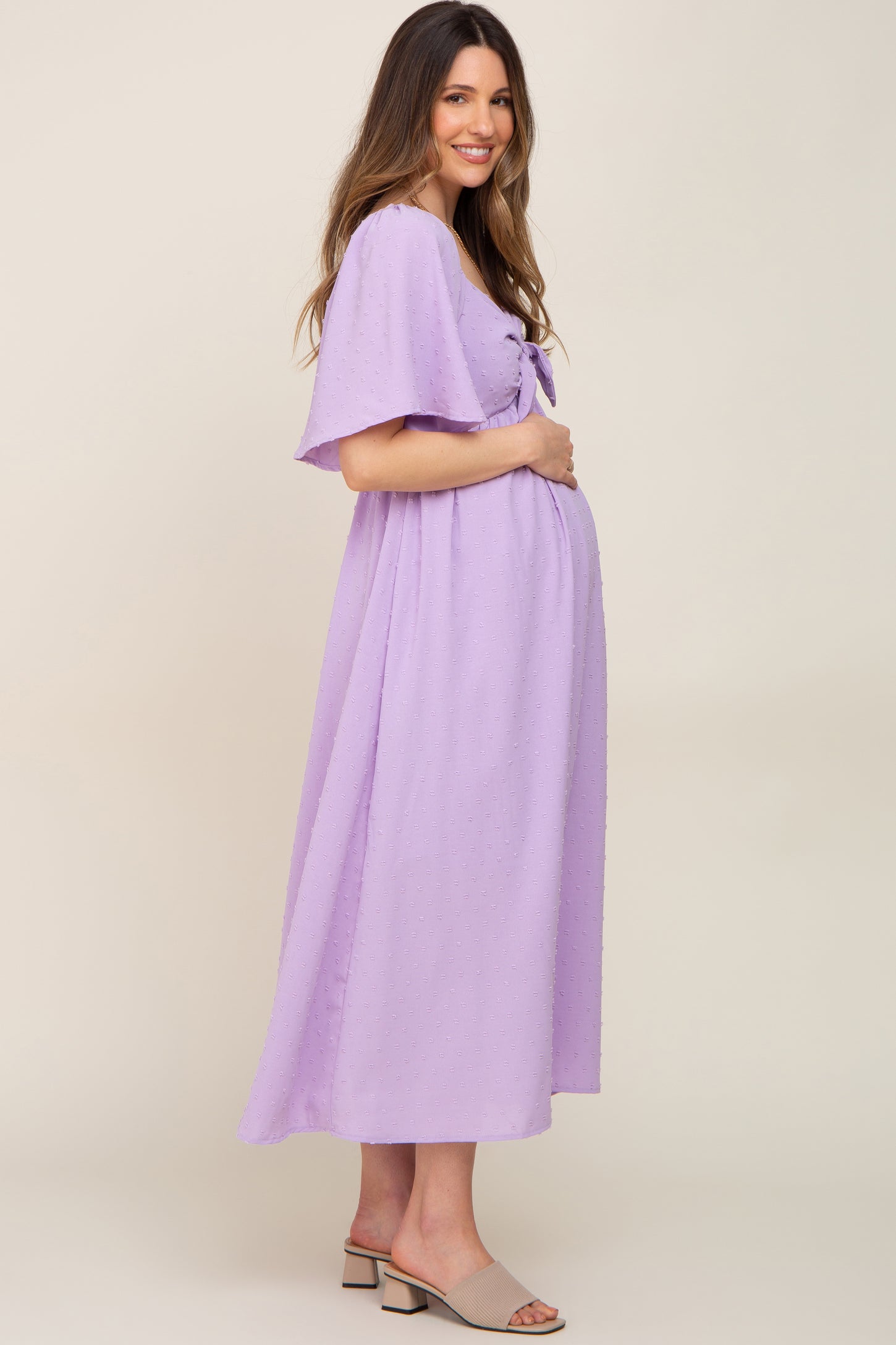 woman wearing a lavender maternity dress with a tie front and flutter sleeves, gently cradling her baby bump