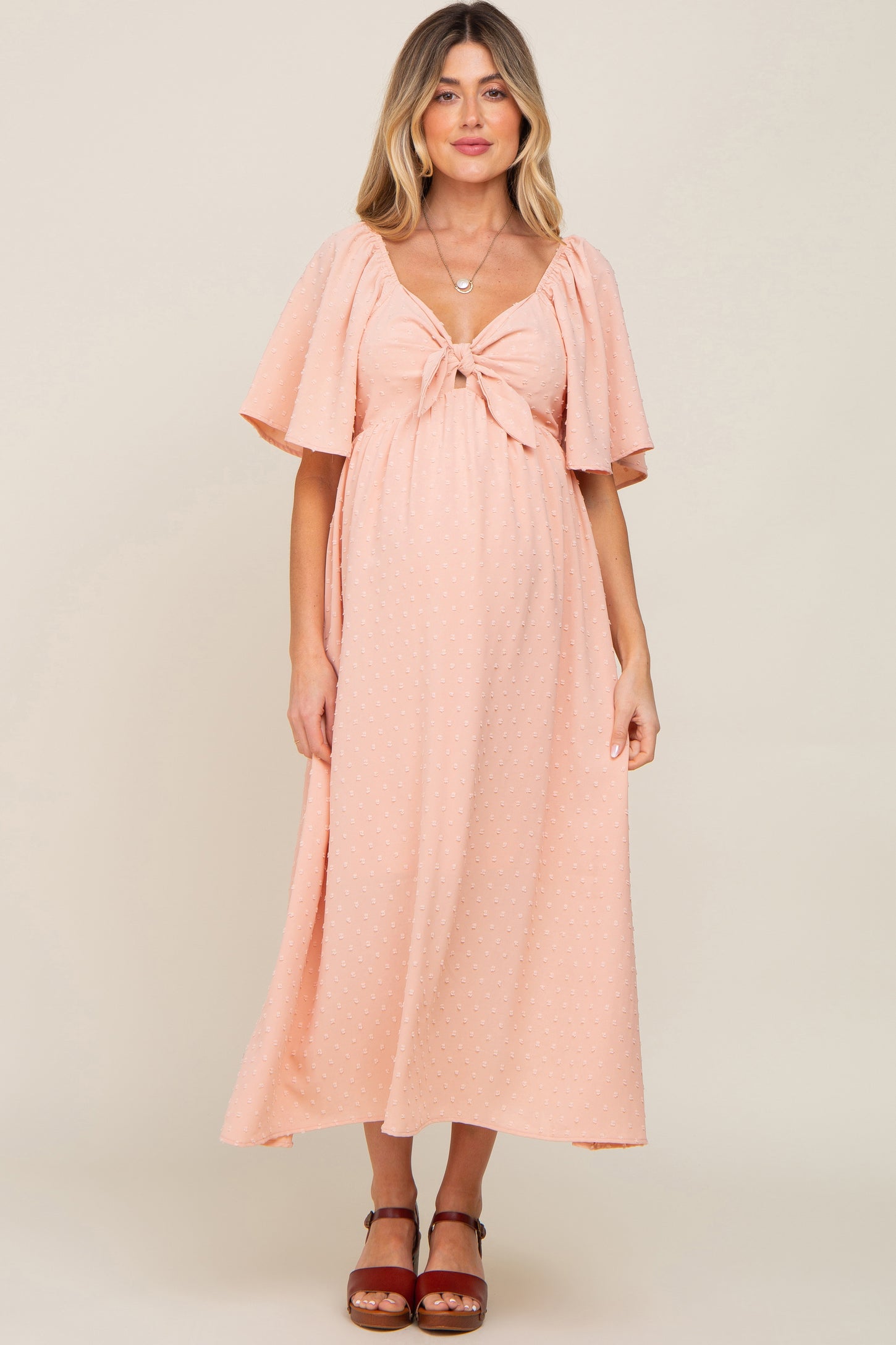 woman wearing a light pink polka dot maternity dress with a bow detail and flutter sleeves, showing her baby bump