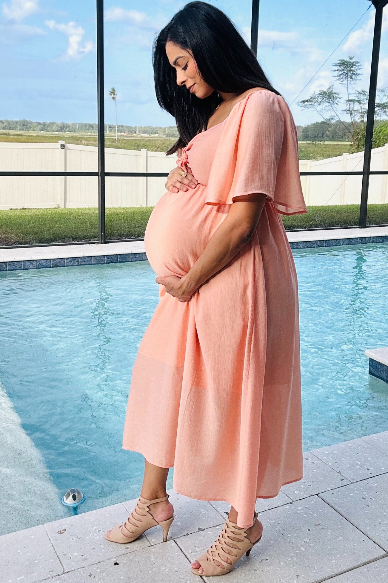 woman wearing a coral maternity dress with short sleeves and a flowy skirt, cradling her baby bump by a pool