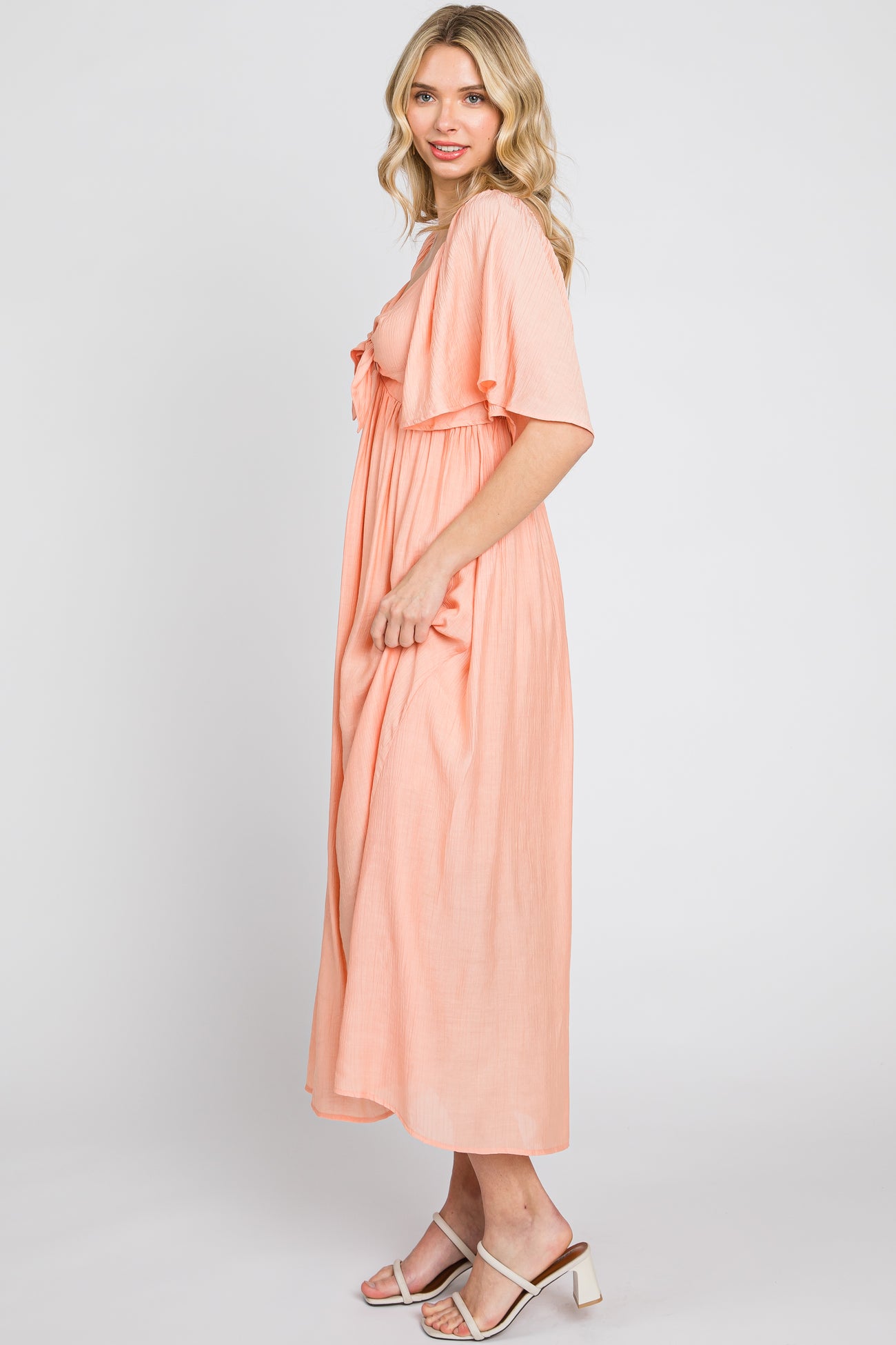 Woman wearing a peach-colored short sleeve maxi dress and white strappy heels against a white background