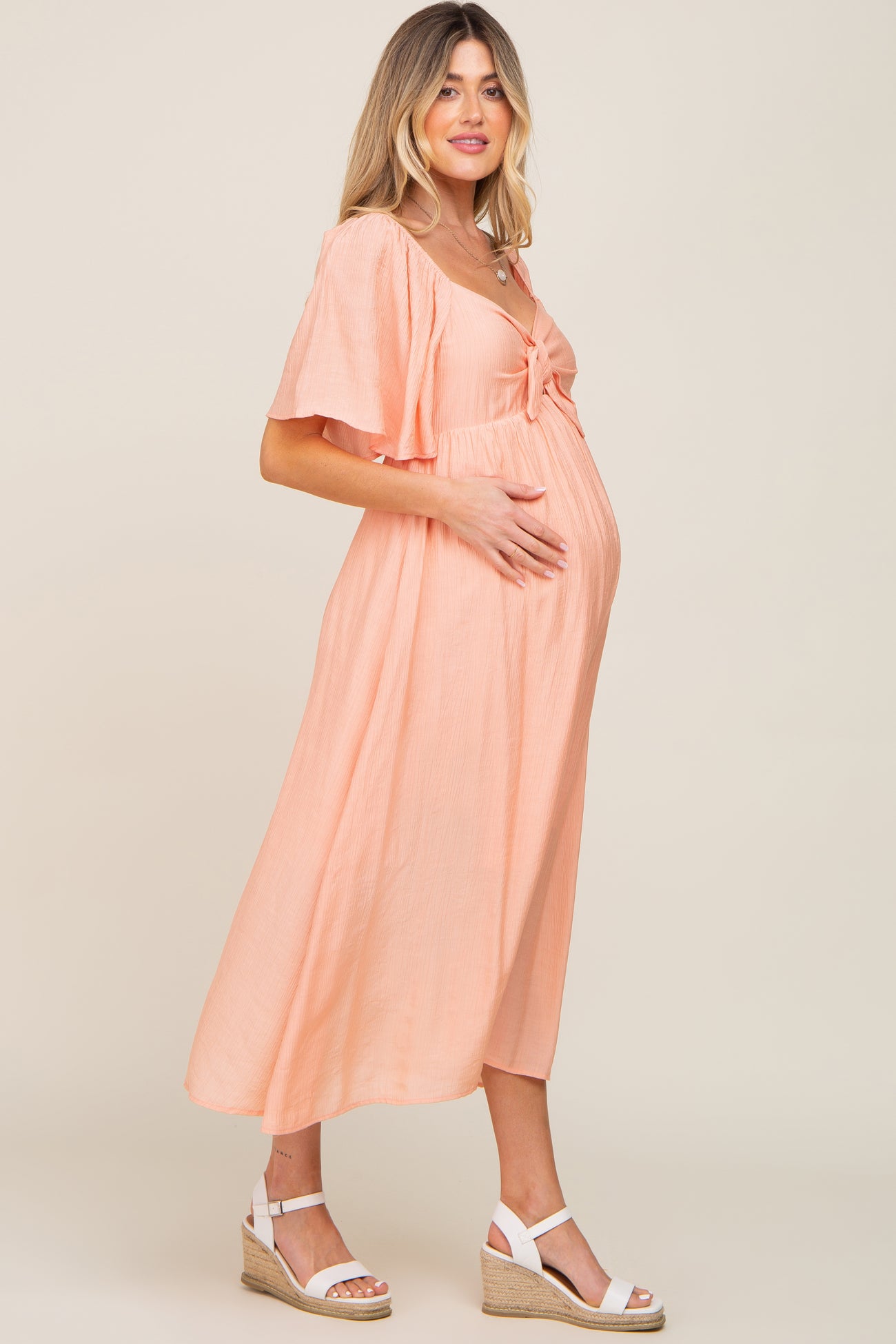 woman wearing a peach maternity dress with flutter sleeves and tie detail, gently holding her baby bump
