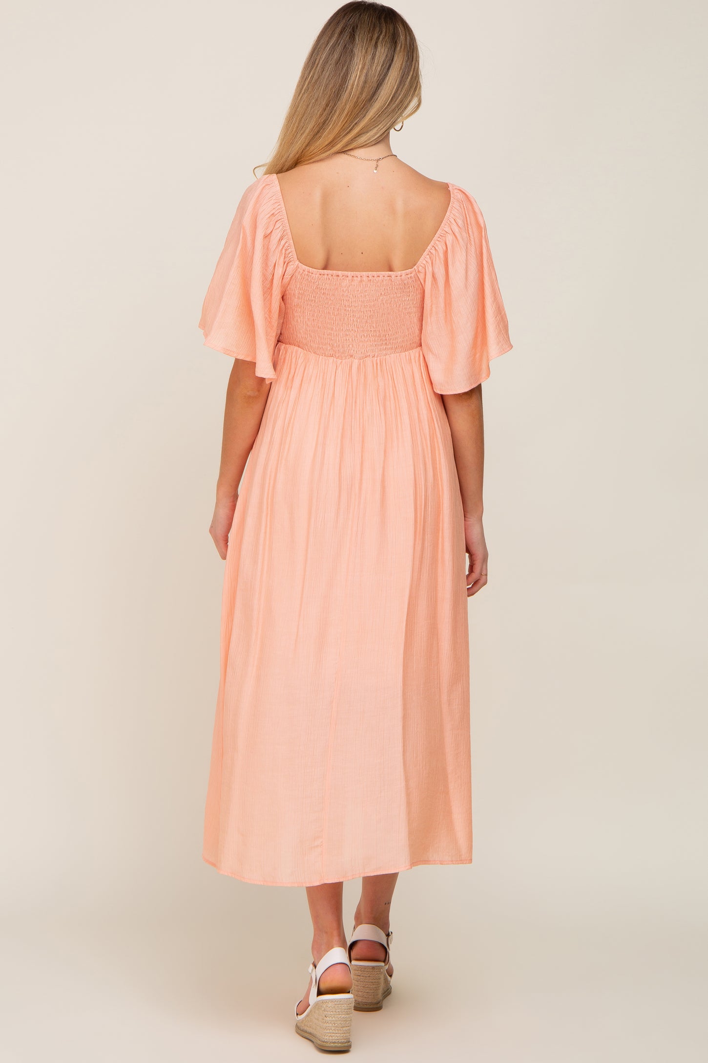 woman wearing a peach maternity dress with flutter sleeves and a smocked back, standing sideways to show the flowy skirt