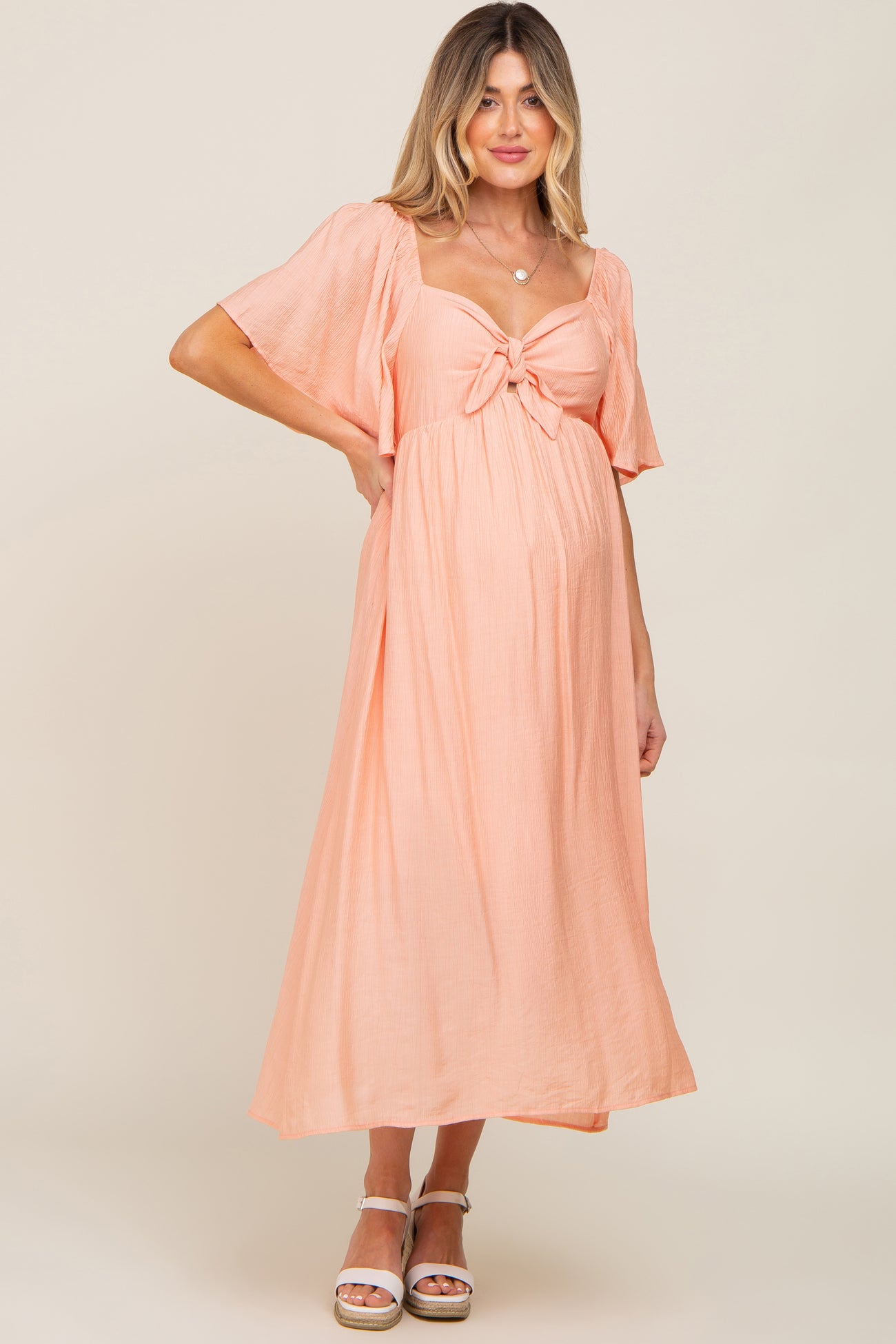 woman wearing a peach maternity maxi dress with a tie detail and flutter sleeves, showing her baby bump