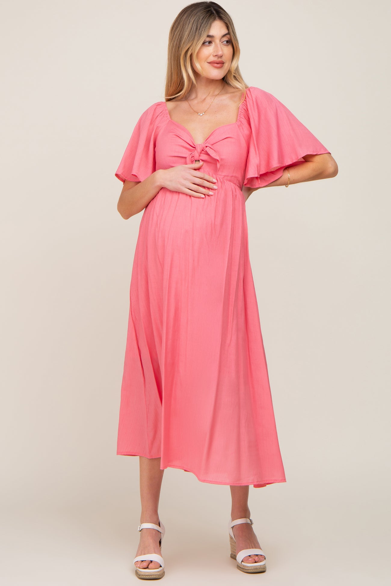 woman wearing a pink maternity dress with flutter sleeves and a tie front, holding her baby bump