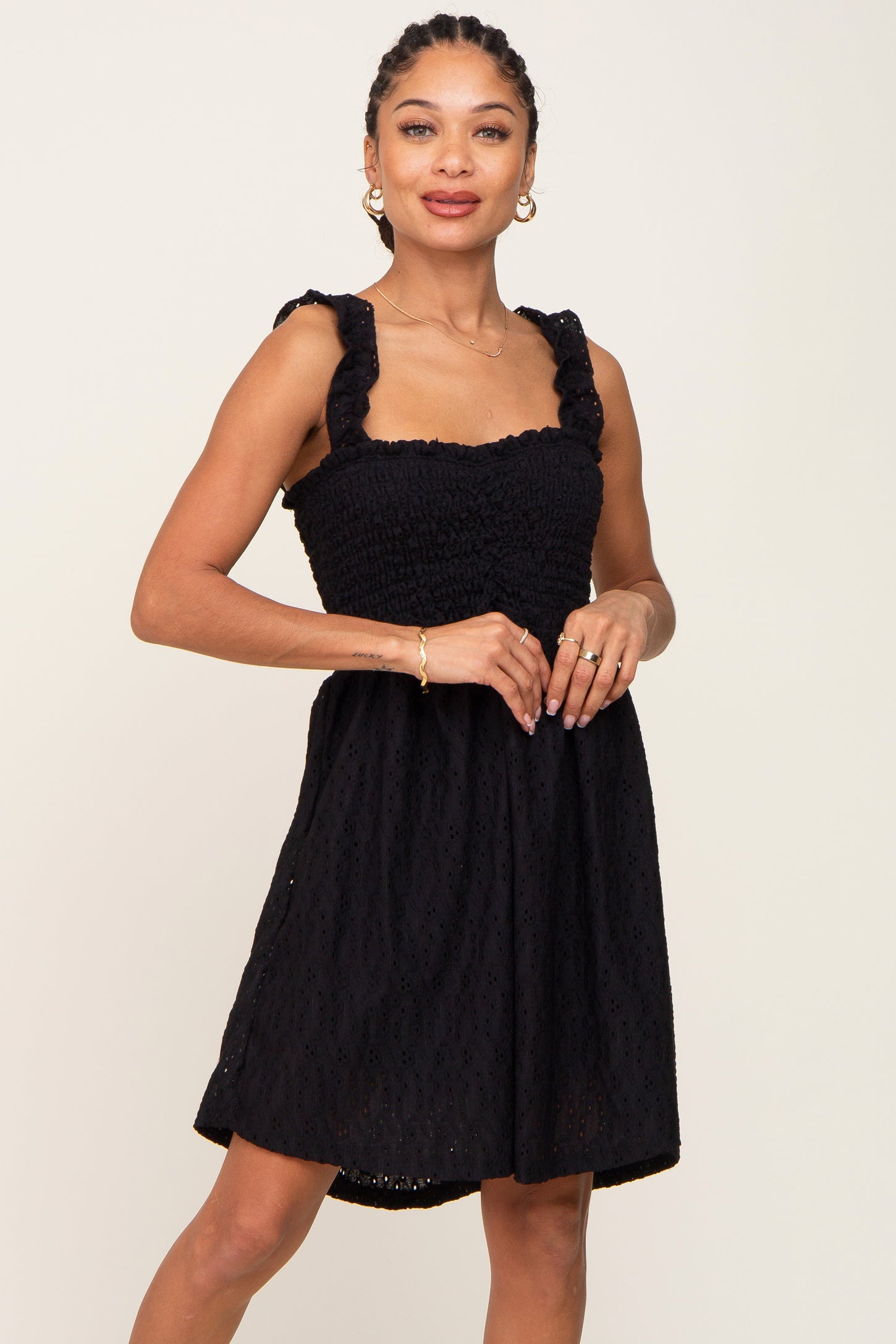 Woman wearing a black smocked eyelet mini dress with ruffle straps and gold jewelry