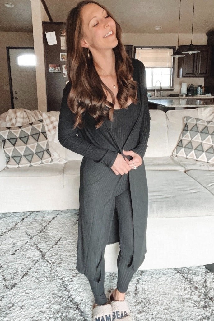 woman wearing a black ribbed jumpsuit with a long cardigan, smiling while standing in a cozy living room