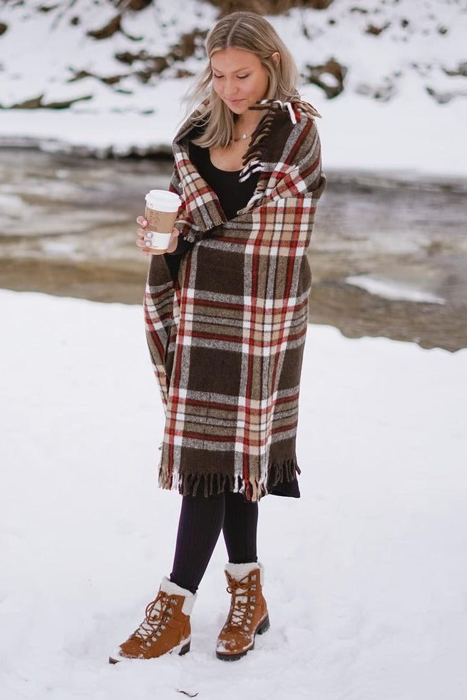 woman wearing a brown plaid wrap shawl, black dress, and brown lace-up boots, holding a coffee cup in a snowy landscape