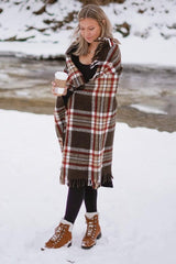 woman wearing a brown plaid wrap shawl, black dress, and brown lace-up boots, holding a coffee cup in a snowy landscape