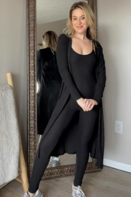 woman wearing a black ribbed long cardigan and matching black bodysuit with leggings, standing in front of a mirror
