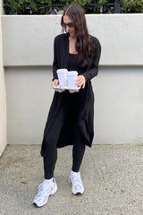 woman wearing a black long cardigan over a black outfit, holding two coffee cups in a tray and wearing white sneakers