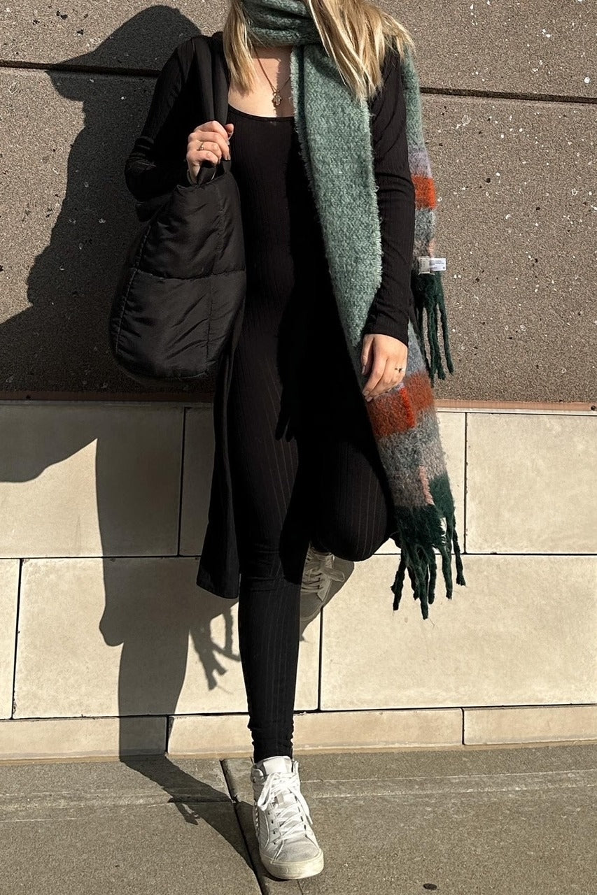 woman wearing a black ribbed jumpsuit with a long black cardigan and a green patterned scarf, holding a black tote bag and wearing sneakers