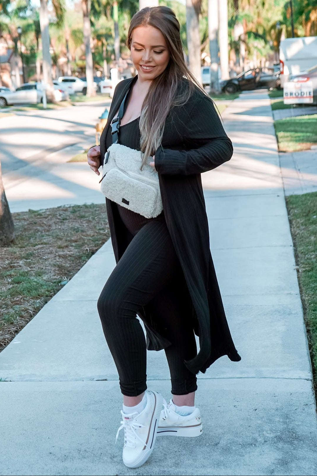 woman wearing a black ribbed long cardigan over a black jumpsuit and white sneakers, carrying a cream crossbody bag