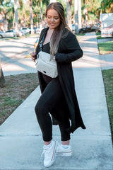 woman wearing a black ribbed long cardigan over a black jumpsuit and white sneakers, carrying a cream crossbody bag