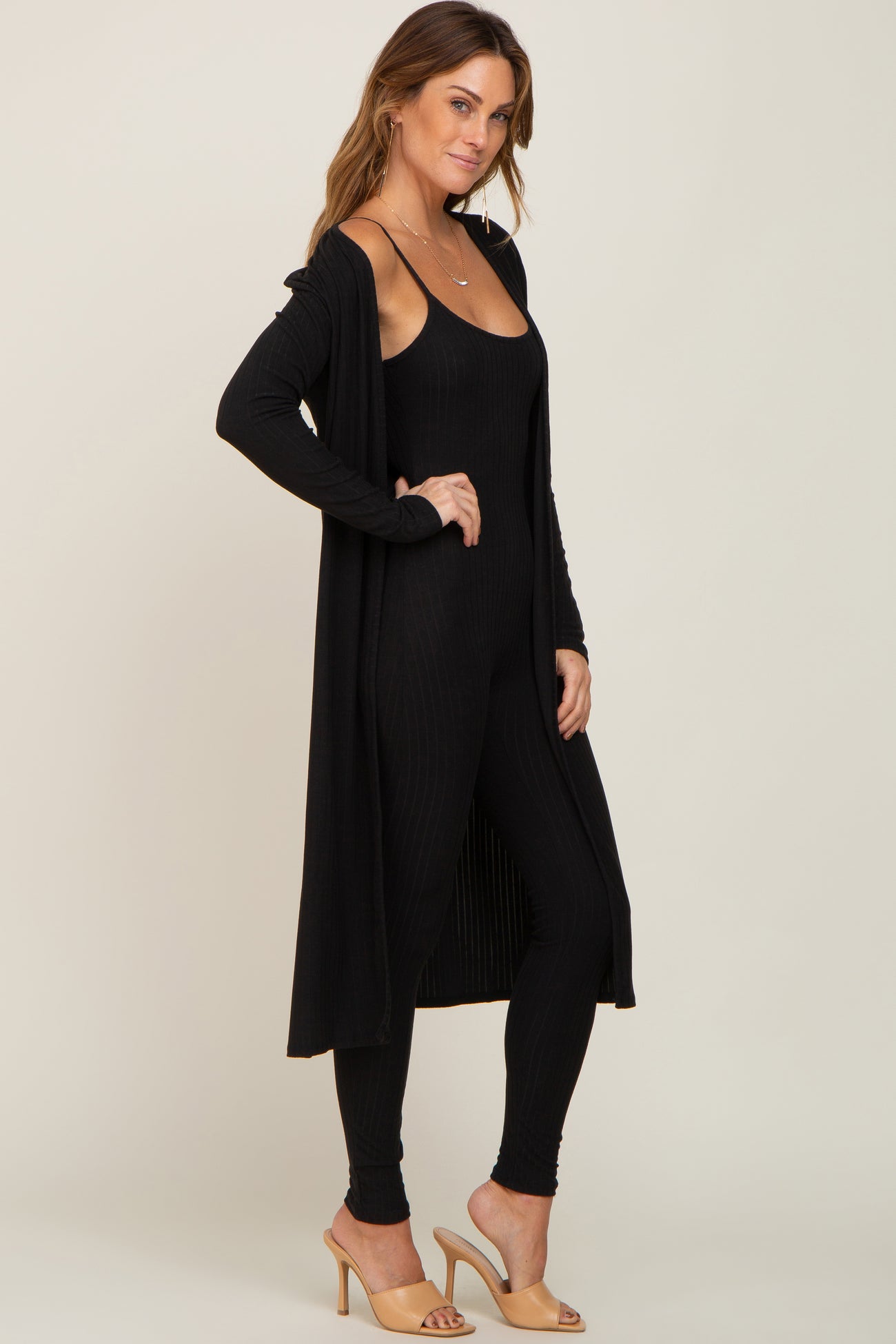 woman wearing a black ribbed long cardigan over a black ribbed jumpsuit and tan heeled mules