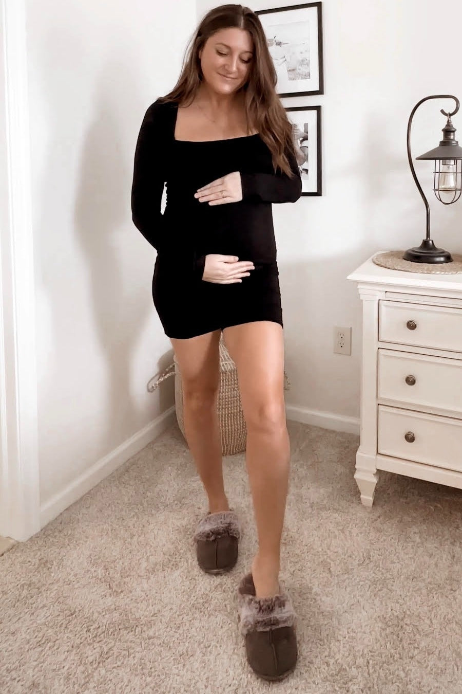 woman wearing a black long sleeve maternity dress and holding her baby bump while standing on carpeted floor