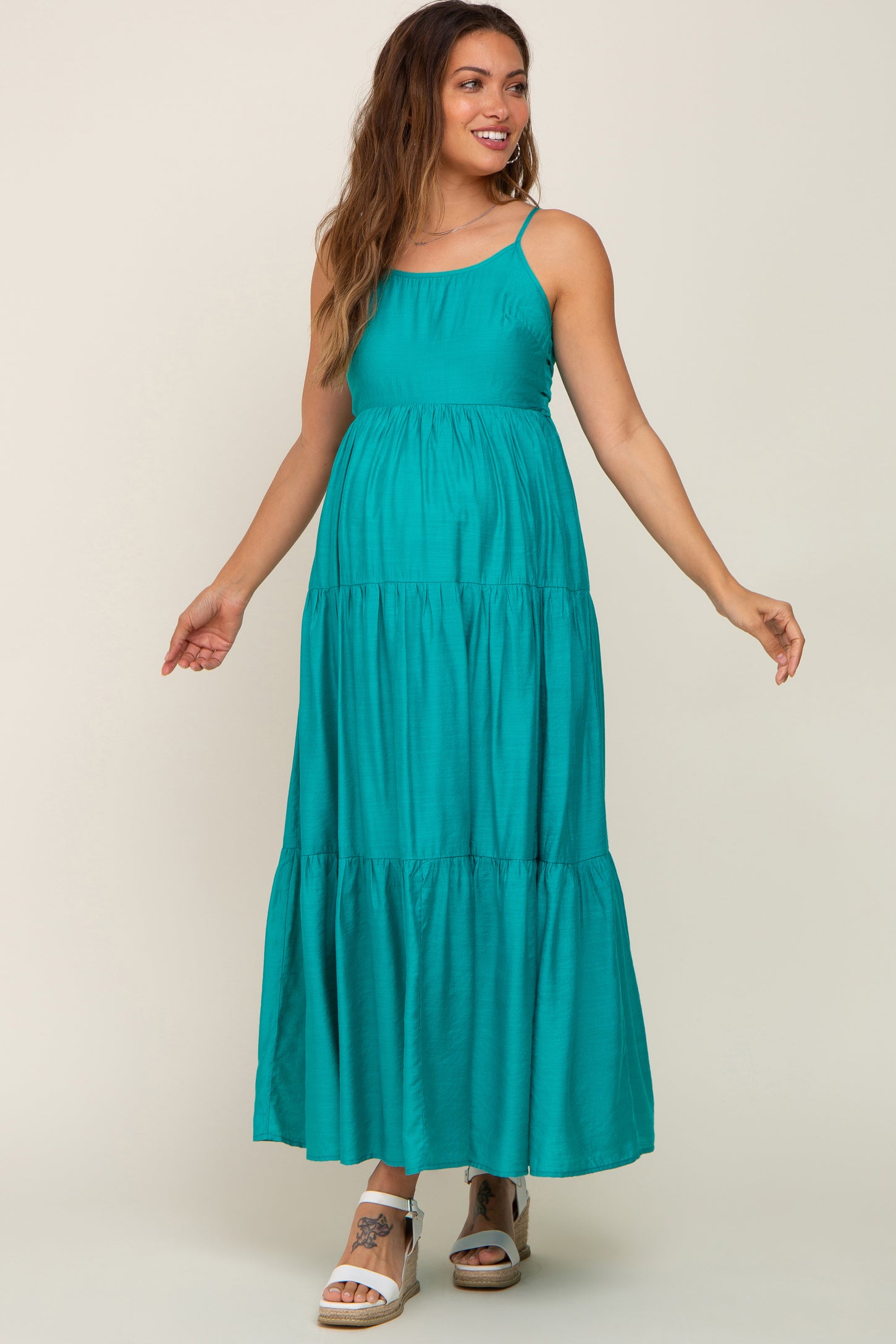 smiling pregnant woman wearing a teal tiered maternity maxi dress with spaghetti straps and white wedge sandals