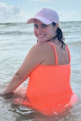 smiling woman in neon orange swimsuit and pink baseball cap sitting in ocean water at the beach