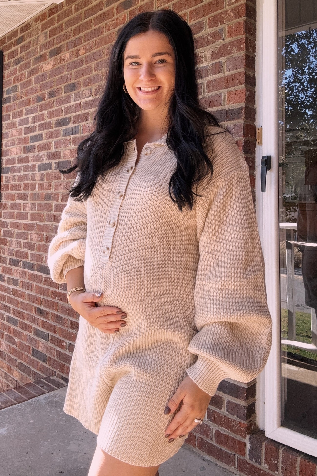 Smiling pregnant woman in a beige knit button-up romper posing in front of a brick wall